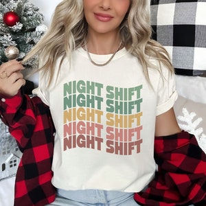 May include: A white t-shirt with the text "NIGHT SHIFT" repeated five times in a rainbow gradient of green, yellow, orange, pink, and red.