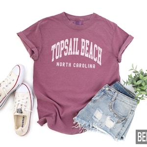 Pu&ograve; includere: Una maglietta color bacca con la scritta "TOPSAIL BEACH NORTH CAROLINA" in bianco. La maglietta &egrave; abbinata a pantaloncini di jeans e scarpe da ginnastica di tela bianche. Un rametto di foglie verdi &egrave; sullo sfondo.