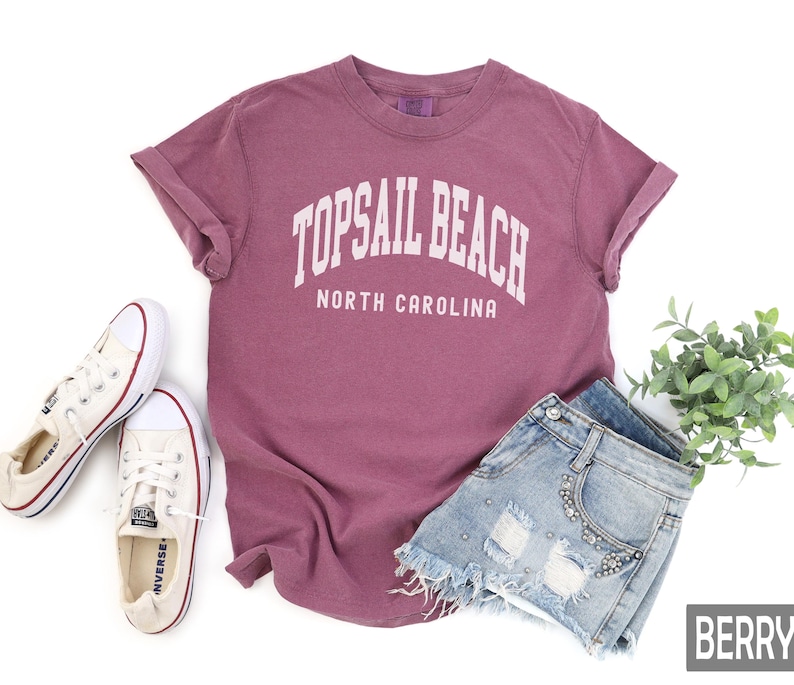Pu&ograve; includere: Una maglietta color bacca con la scritta "TOPSAIL BEACH NORTH CAROLINA" in bianco. La maglietta &egrave; abbinata a pantaloncini di jeans e scarpe da ginnastica di tela bianche. Un rametto di foglie verdi &egrave; sullo sfondo.