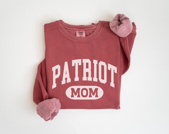 May include: A maroon sweatshirt with the words "PATRIOT MOM" in white block letters. The sweatshirt has a crew neck and long sleeves, with a matching scrunchie on the sleeve. The background is white.
