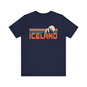 Puede incluir: Una camiseta azul marino con un gr&aacute;fico retro de monta&ntilde;as y un sol con la palabra "ICELAND" en texto naranja.