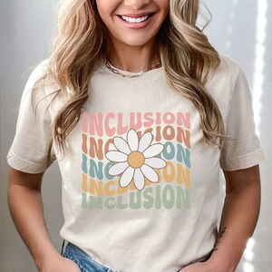 May include: A beige t-shirt with a daisy graphic and the word "INCLUSION" repeated in a rainbow pattern.