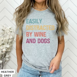 May include: A heather grey t-shirt with the text "EASILY DISTRACTED BY WINE AND DOGS" printed in colorful retro-style lettering.