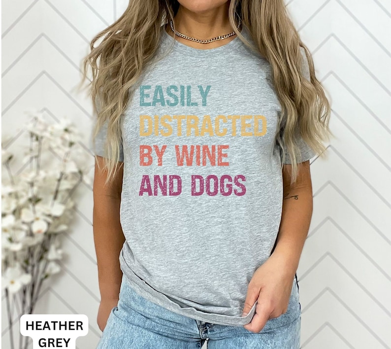 May include: A heather grey t-shirt with the text "EASILY DISTRACTED BY WINE AND DOGS" printed in colorful retro-style lettering.