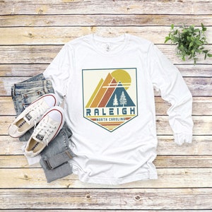 May include: White long-sleeve t-shirt with a vintage-style graphic design featuring a mountain range, trees, and a sun. The graphic is in a shield shape with the text "Raleigh, North Carolina" below it.