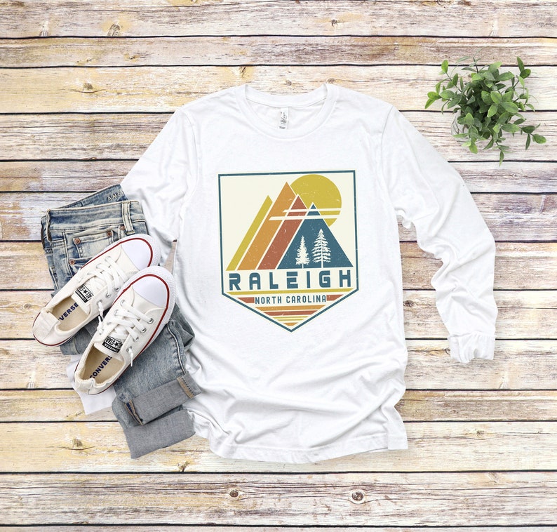 May include: White long-sleeve t-shirt with a vintage-style graphic design featuring a mountain range, trees, and a sun. The graphic is in a shield shape with the text "Raleigh, North Carolina" below it.