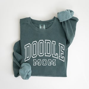Peut inclure: Un sweat-shirt bleu sarcelle avec le texte "DOODLE MOM" en lettres blanches soulignées.