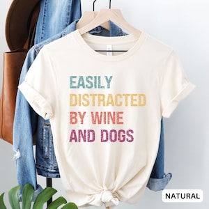 May include: A white t-shirt with the text "EASILY DISTRACTED BY WINE AND DOGS" in a vintage style font with rainbow colors. The shirt is on a hanger and has a natural color.