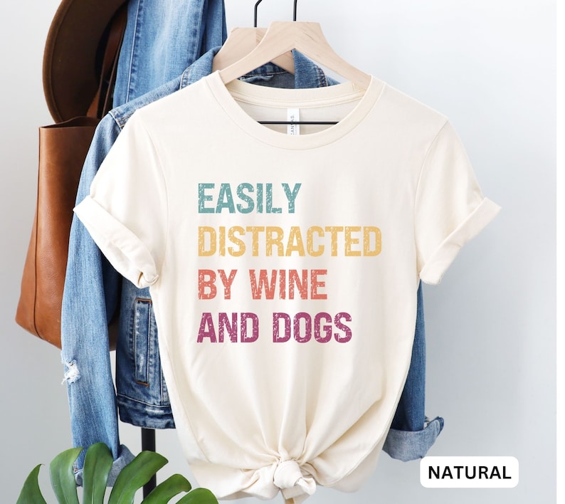 May include: A white t-shirt with the text "EASILY DISTRACTED BY WINE AND DOGS" in a vintage style font with rainbow colors. The shirt is on a hanger and has a natural color.