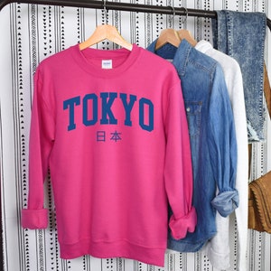 以下が含まれることがあります： ピンクのトレーナーに、紺色の文字で「TOKYO」と、その下に紺色の小さな文字で日本の日本語「日本」が書かれています。