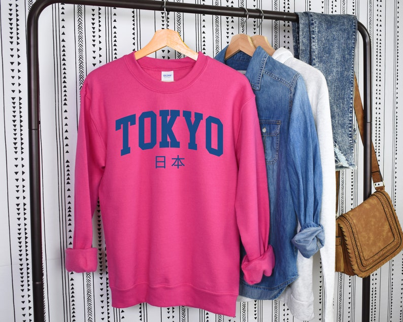 以下が含まれることがあります： ピンクのトレーナーに、紺色の文字で「TOKYO」と、その下に紺色の小さな文字で日本の日本語「日本」が書かれています。