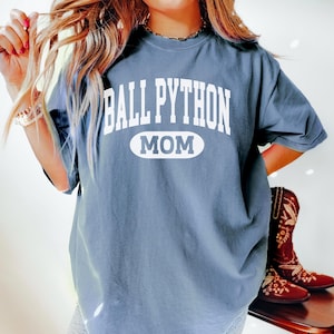 Puede incluir: Una camiseta azul grisáceo con el texto blanco "BALL PYTHON MOM". La palabra "MOM" está en un óvalo blanco. La camiseta es de estilo casual, adecuada para el uso diario.