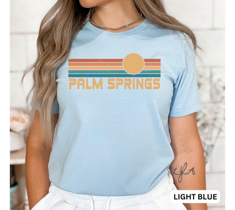 May include: Light blue t-shirt with a retro-style graphic of a sunset over a striped horizon and the text "PALM SPRINGS".