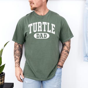 May include: Green short-sleeved t-shirt with the text "TURTLE DAD" in white block letters. The text is centered on the chest. The shirt is made of a soft, comfortable material.