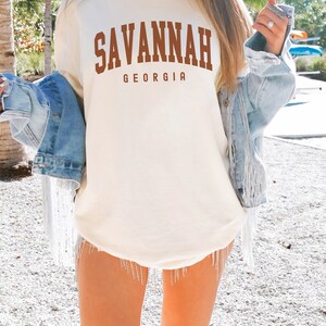 Op de afbeelding: Ivoor kleurig t-shirt met het woord "SAVANNAH" in bruine blokletters, met "GEORGIA" eronder. Het model draagt een spijkerjasje en shorts met strass-franjes.