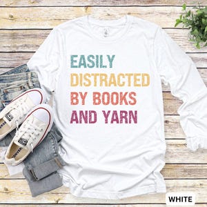 May include: White long-sleeve t-shirt with the text "EASILY DISTRACTED BY BOOKS AND YARN" in a rainbow color scheme.
