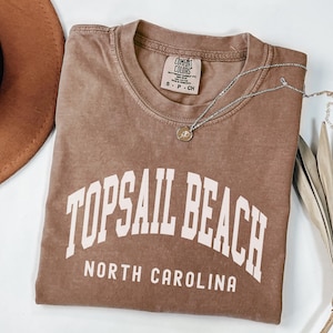 Pu&ograve; includere: Una maglietta marrone con la scritta "TOPSAIL BEACH NORTH CAROLINA" a blocchi bianchi. Una collana d'argento con un piccolo ciondolo &egrave; appoggiata sulla maglietta. Un cappello marrone &egrave; visibile nell'angolo in alto a sinistra.