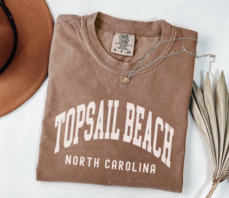 Pu&ograve; includere: Una maglietta marrone con la scritta "TOPSAIL BEACH NORTH CAROLINA" a blocchi bianchi. Una collana d'argento con un piccolo ciondolo &egrave; appoggiata sulla maglietta. Un cappello marrone &egrave; visibile nell'angolo in alto a sinistra.
