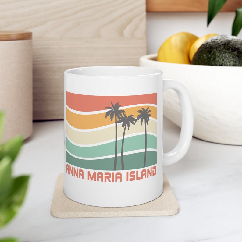 Puede incluir: Taza de cer&aacute;mica blanca con un dise&ntilde;o retro que presenta palmeras y el texto "Anna Maria Island". La taza tiene un asa y est&aacute; sobre un posavasos beige.