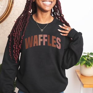 May include: A black sweatshirt with the word "WAFFLES" printed in brown letters across the front. The letters are in a gradient of brown shades.