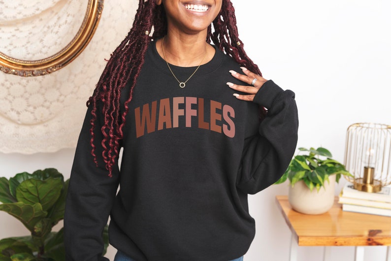 May include: A black sweatshirt with the word "WAFFLES" printed in brown letters across the front. The letters are in a gradient of brown shades.