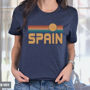 May include: A heather navy blue t-shirt with a vintage-style graphic of a sunset over a striped horizon with the word "SPAIN" in a retro font.