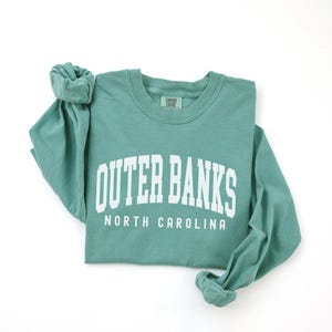 May include: A teal long-sleeve shirt with the words "OUTER BANKS" in large white block letters, and "NORTH CAROLINA" below. The shirt is folded, with the sleeves rolled up.