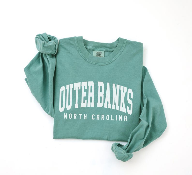 May include: A teal long-sleeve shirt with the words "OUTER BANKS" in large white block letters, and "NORTH CAROLINA" below. The shirt is folded, with the sleeves rolled up.