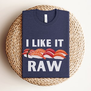 以下が含まれることがあります： 白地に「I like it RAW」と書いてある紺色のTシャツ。Tシャツには、さまざまな具材の入った寿司ロール5つのグラフィックが描かれている。