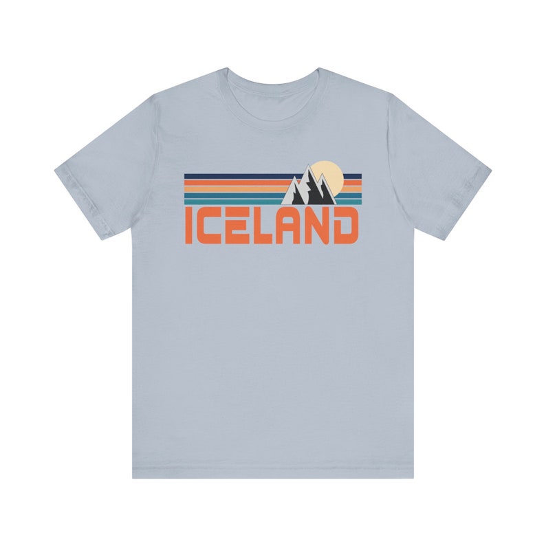 Puede incluir: Una camiseta azul claro con un gr&aacute;fico retro de monta&ntilde;as, un sol y la palabra "ICELAND" en naranja.