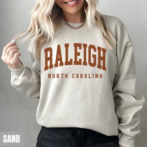 Pode incluir: Uma camisa de moletom bege com o texto "RALEIGH NORTH CAROLINA" em letras marrons.
