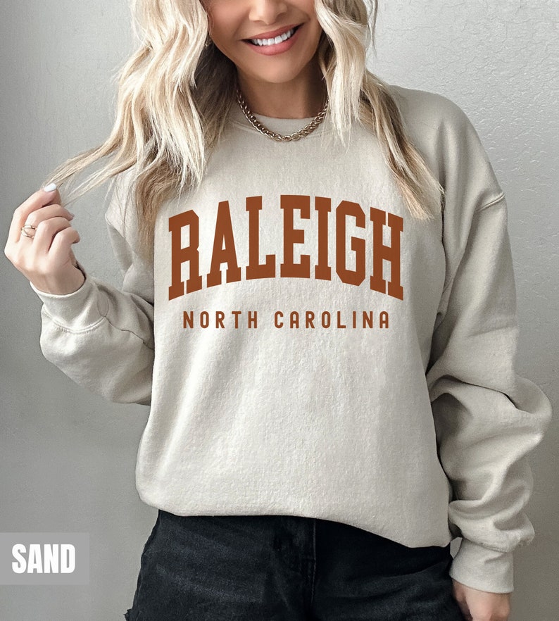Pode incluir: Uma camisa de moletom bege com o texto "RALEIGH NORTH CAROLINA" em letras marrons.