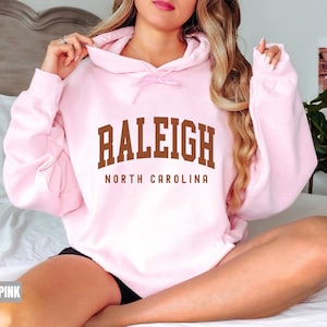 Pode incluir: Um moletom rosa claro com o texto "Raleigh North Carolina" em letras marrons.