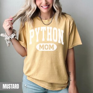 May include: Mustard-coloured t-shirt with the text "PYTHON MOM" in white block letters. The word "MOM" is in an oval shape. The shirt has short sleeves and a relaxed fit. The shirt is made of a soft, comfortable material.