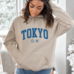 以下が含まれることがあります： ベージュのフード付きスウェットシャツに、青い「TOKYO」のグラフィックと、日本の日本語「日本」が下に印刷されています。