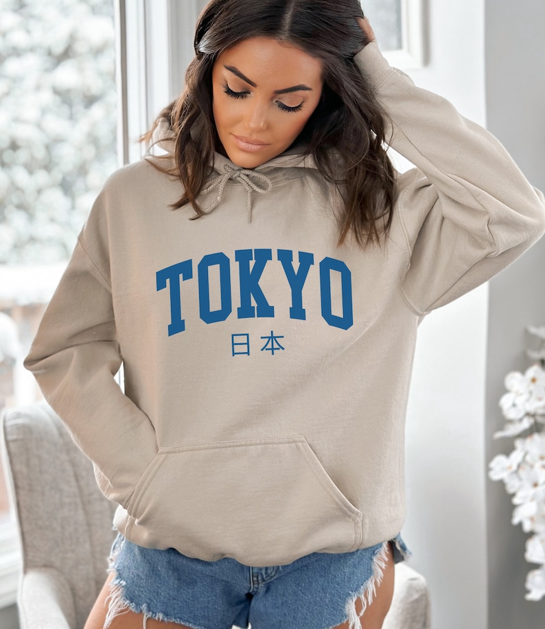 以下が含まれることがあります： ベージュのフード付きスウェットシャツに、青い「TOKYO」のグラフィックと、日本の日本語「日本」が下に印刷されています。