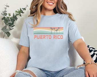 May include: A light blue t-shirt with a retro-style graphic of palm trees and a sunset. The text "PUERTO RICO" is printed in red below the graphic.