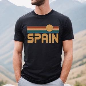 May include: A black t-shirt with a vintage-style graphic design featuring a sunset with a yellow sun and the word "SPAIN" in a retro font.