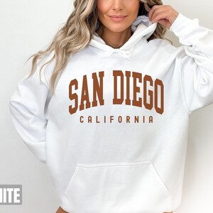 Pode incluir: Moletom branco com capuz com uma estampa marrom que diz "San Diego California".
