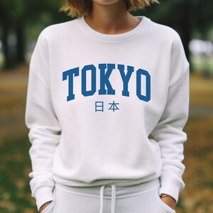 以下が含まれることがあります： 白いスウェットシャツに青い「TOKYO」のグラフィックと、その下に日本語で「日本」と印刷されています。