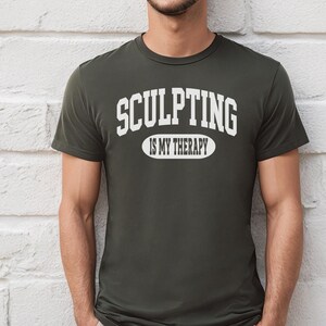 K&ouml;nnte beinhalten: Dunkel olivgr&uuml;nes T-Shirt mit dem Text "SCULPTING IS MY THERAPY" in wei&szlig;en Blockbuchstaben. Der Text ist bogenf&ouml;rmig &uuml;ber einem Oval angeordnet. Das Shirt hat kurze &Auml;rmel.