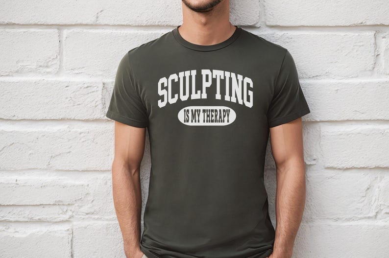 K&ouml;nnte beinhalten: Dunkel olivgr&uuml;nes T-Shirt mit dem Text "SCULPTING IS MY THERAPY" in wei&szlig;en Blockbuchstaben. Der Text ist bogenf&ouml;rmig &uuml;ber einem Oval angeordnet. Das Shirt hat kurze &Auml;rmel.