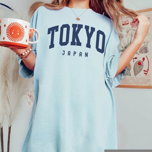 Pode incluir: Uma camiseta azul claro oversized com o texto "TOKYO JAPAN" em letras azul marinho. A camiseta &eacute; feita de um tecido chambray macio e confort&aacute;vel.