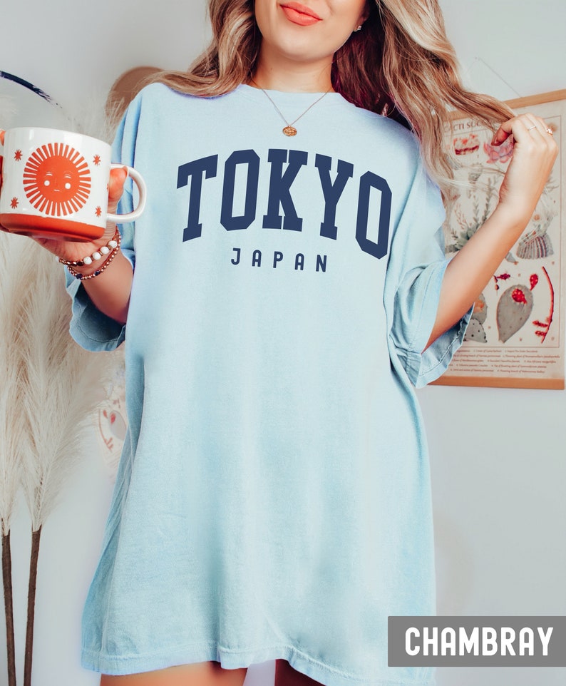 Pode incluir: Uma camiseta azul claro oversized com o texto "TOKYO JAPAN" em letras azul marinho. A camiseta &eacute; feita de um tecido chambray macio e confort&aacute;vel.