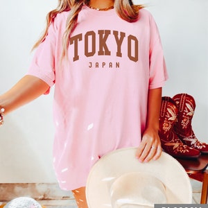 Pode incluir: Uma camiseta rosa oversized com o texto "TOKYO JAPAN" em letras marrons. A camiseta est&aacute; sendo usada por uma pessoa com um chap&eacute;u branco e cal&ccedil;as marrons.
