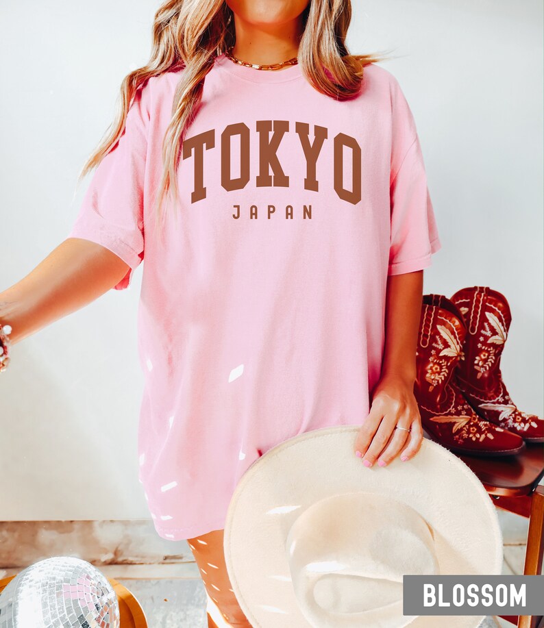 Pode incluir: Uma camiseta rosa oversized com o texto "TOKYO JAPAN" em letras marrons. A camiseta est&aacute; sendo usada por uma pessoa com um chap&eacute;u branco e cal&ccedil;as marrons.