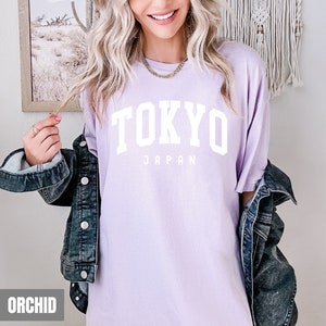 Pode incluir: Uma camiseta lavanda com o texto "TOKYO JAPAN" em letras brancas.
