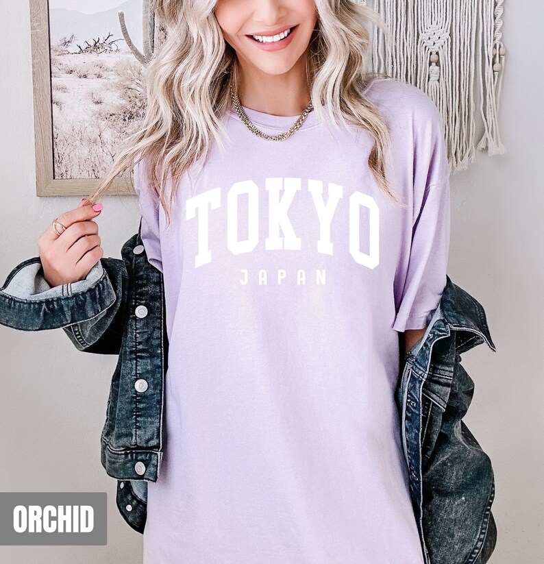 Pode incluir: Uma camiseta lavanda com o texto "TOKYO JAPAN" em letras brancas.