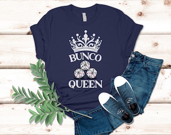 May include: Navy blue t-shirt with a white crown design, the text "Bunco Queen", and dice with pink hearts. The shirt is paired with blue jeans and sneakers, arranged on a wooden surface.