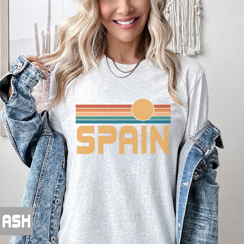 May include: A light grey t-shirt with a retro-style graphic design featuring a sunset with a sun and the word "SPAIN" in orange.